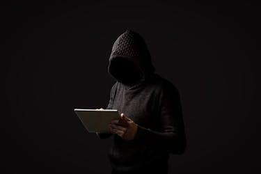Faceless man in a hoodie with a hood holds a tablet in his hands on a dark background. Concept of hacking user data. Hacked lock, credit card, cloud, email, passwords, personal files